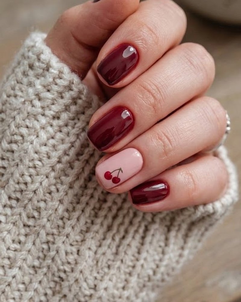 Esmaltado semipermanente en color vino sobre uñas cortas, con un diseño de cerezas en una uña